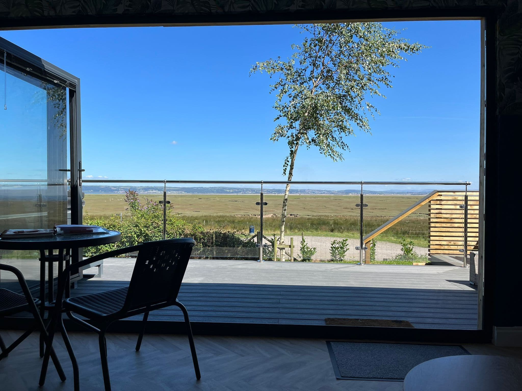 A café table and two chairs on the Langland Bay Lookout's deck on a clear day, a young birch tree and the flat marsh beyond the rail