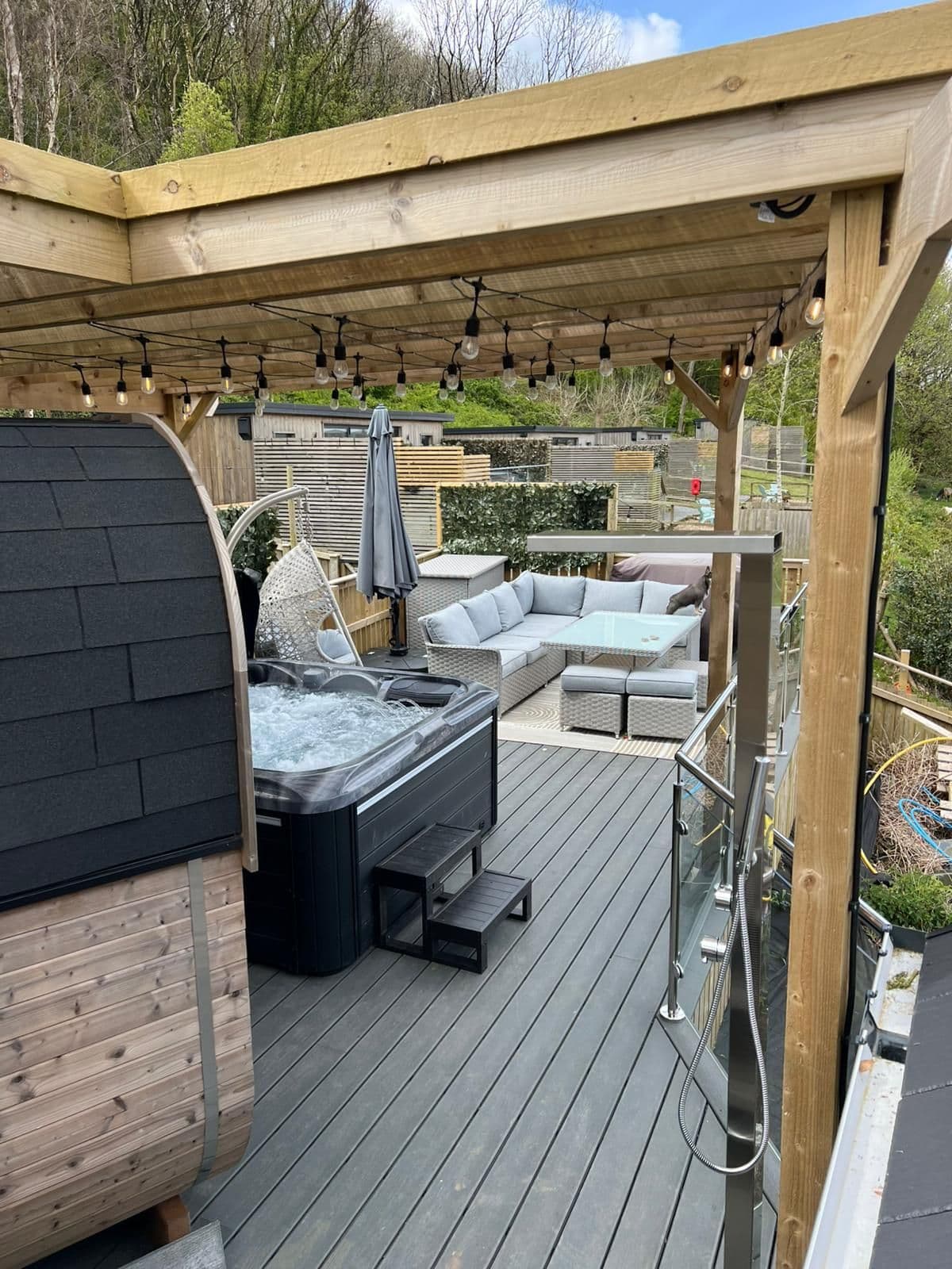 Festoon lights strung across the timber pergola at the Oxwich Bay Lodge, over corner seating, a hanging chair and the hot tub