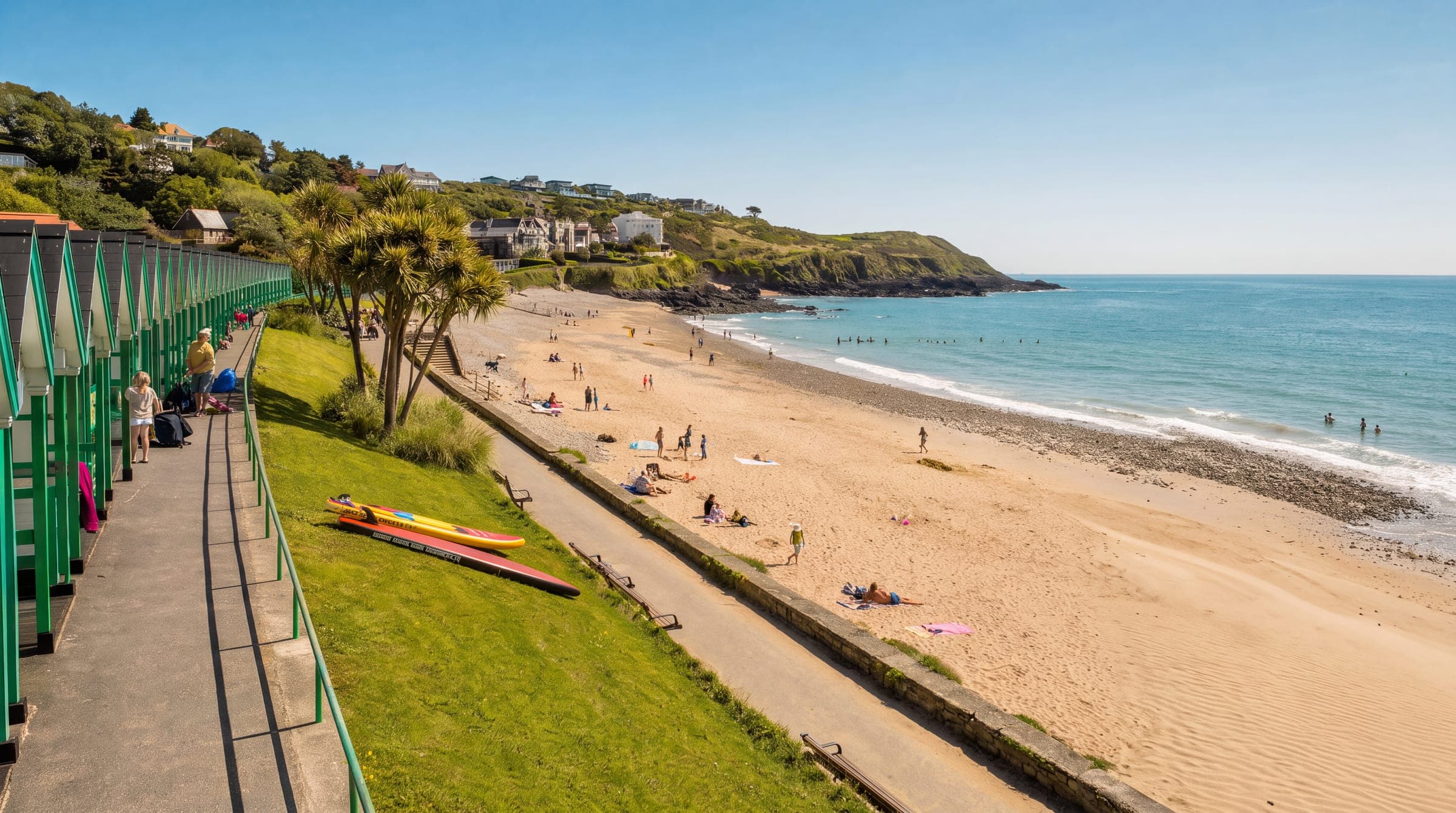 Langland Bay from the clifftop path: the row of green-and-white beach huts and the promenade on the left, palms on the grass bank, a broad sweep of sand with bathers in the shallows, and the headland closing the bay on the right