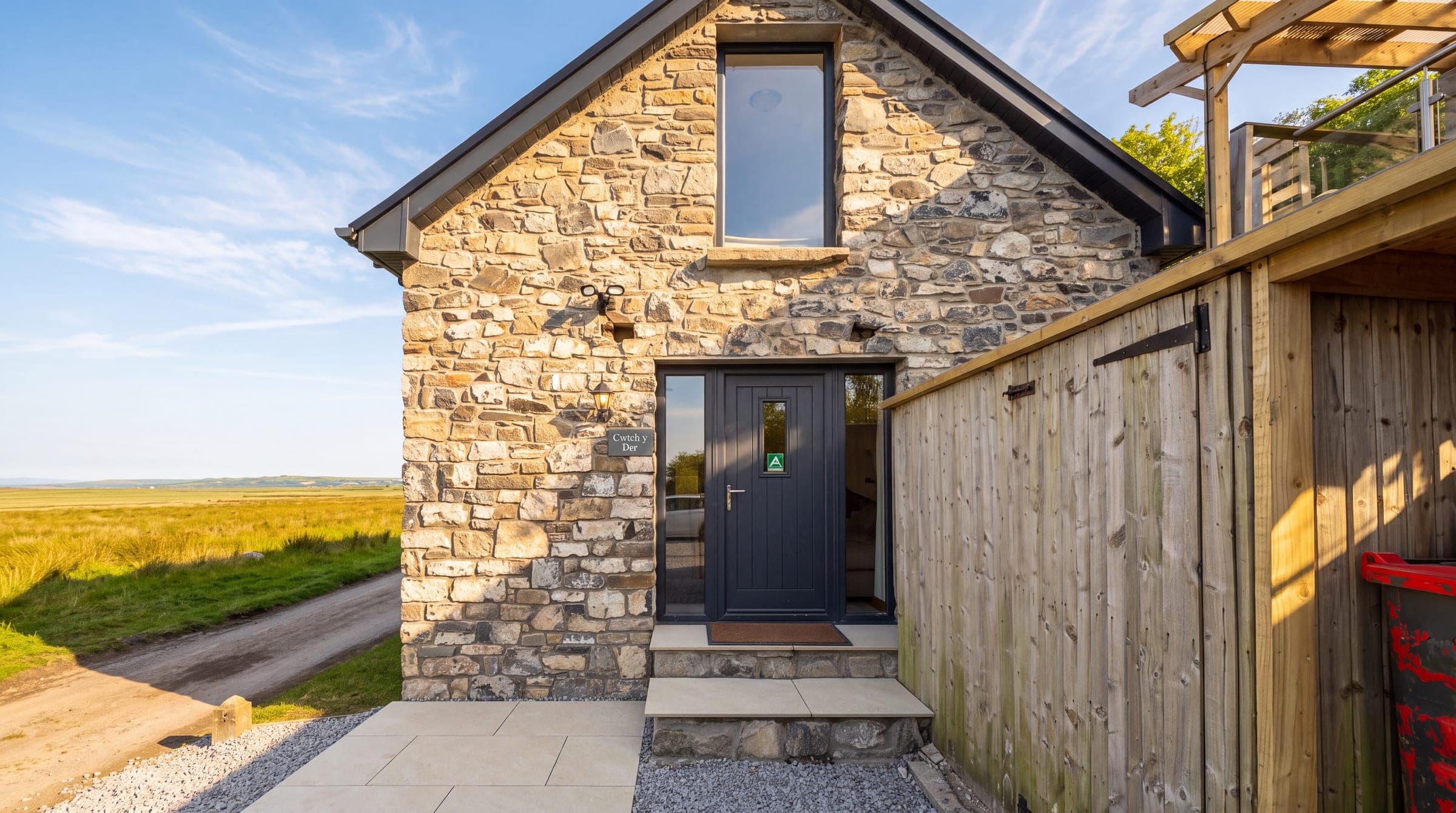 The stone gable end of the Oxwich Bay Lodge in low evening light, its dark front door open at the top of two stone steps beside a small slate name plaque, a timber store to the right and the flat marsh stretching away on the left