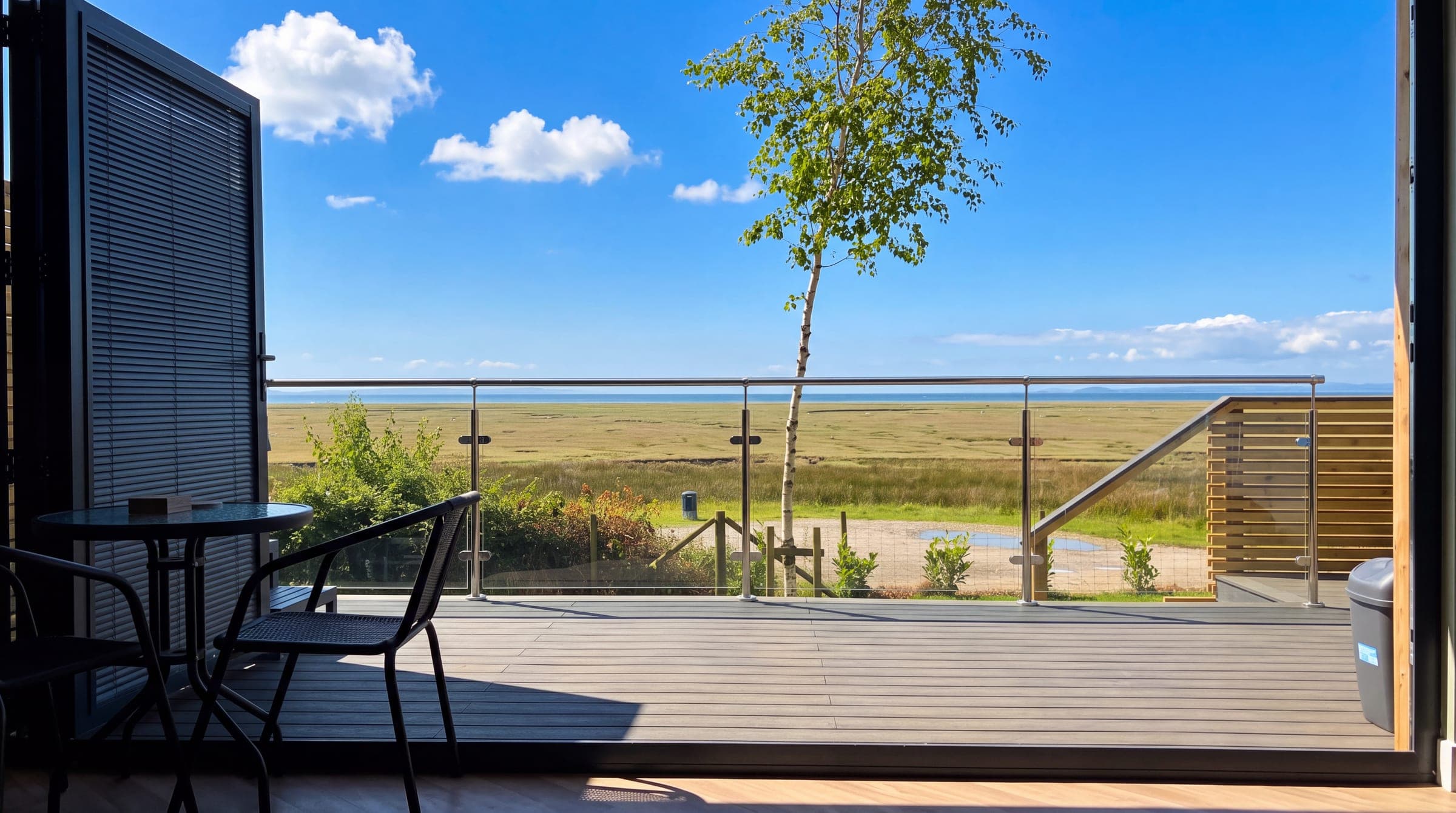 Looking out from inside a cabin across its decking: a bistro table and chairs to the left, a young silver birch beyond the glass balustrade, and the flat green salt marsh running out to the estuary and a blue sky